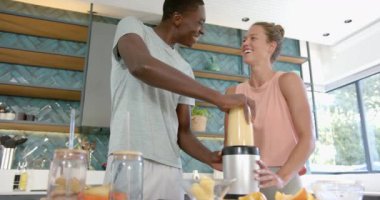 Çeşitli çiftler mutfakta, evde smoothie yapıyorlar. İkisi de gülümsüyor, beyaz bir kadın Afrikalı bir Amerikalının eline rehberlik ediyor, değişmeden, ağır çekimde.