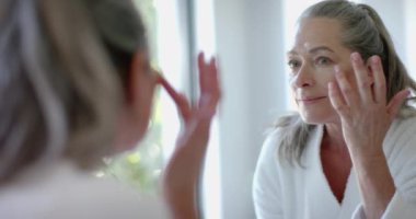 Cilt bakımı, bornozlu olgun bir kadın aynaya bakıyor, gülümsüyor. Güzellik, kendine dikkat, sabah rutini, tazelik, değişmemiş, yavaş çekim