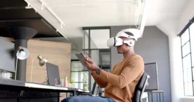 VR kulaklığı kullanarak, Afrikalı Amerikalı bir adam modern ofis çalışma alanındaki tabletle etkileşime geçiyor. Teknoloji, sanal gerçeklik, aygıt, dijital, yenilik, iş