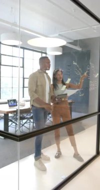 Dikey video: Ofiste işbirliği yapmak, meslektaşların cam tahtaya yazı yazıp beyin fırtınası yapması. Takım çalışması, işbirliği, toplantı, strateji, planlama, yenilik