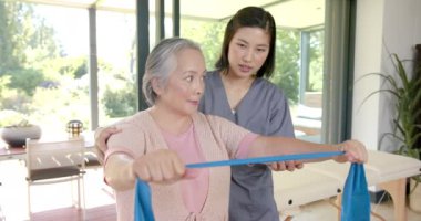 Kıdemli Asyalı kadın direniş bandıyla egzersiz yapıyor, evde fizyoterapistin yardımıyla. Egzersiz, fitness, rehabilitasyon, sağlık, eğitim, terapi