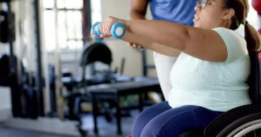 Tekerlekli sandalyedeki kadın, halterlerle egzersiz yapıyor, spor salonunda terapistin de yardımıyla. Fitness, egzersiz, rehabilitasyon, fizyoterapi, sağlık, güç, değişmemiş, yavaş çekim
