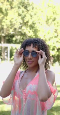 Dikey video: Gülümseyen kadın güneş gözlüğü tutuyor, açık havada güneşli bir günün keyfini çıkarıyor. yaz, mutluluk, moda, eğlence, doğa, yaşam tarzı