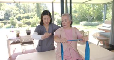 Evde fizyoterapist rehberliğinde direniş bandıyla egzersiz yapan kıdemli Asyalı kadın. Egzersiz, fitness, rehabilitasyon, terapi, sağlık, refah