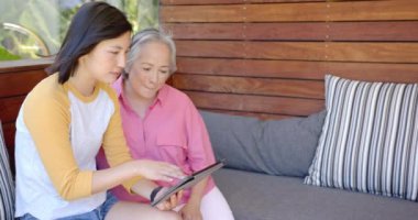 Tablet kullanarak, genç Asyalı kadın kanepedeki yaşlı Asyalı kadına bilgi veriyor. Teknoloji, akıl hocalığı, öğrenme, öğretim, bağlanma, etkileşim