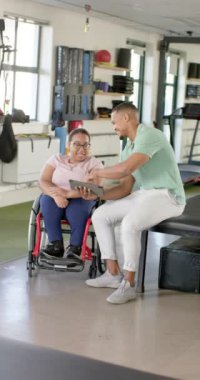 Dikey video: Engelli rehabilitasyonunda, tekerlekli sandalyedeki kadına tablet gösteriliyor, terapist ilerleme tartışıyor. Fizyoterapi, rehabilitasyon, sağlık, hasta, teknoloji, iyileşme, yavaş çekim