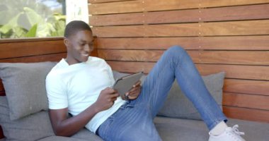 Kanepede dinlenirken, Afro-Amerikan adam gülümsüyor ve eğlence için tablet kullanıyor. Rahatlama, rahatlama, teknoloji, tarama, elektronik cihaz, konforlu