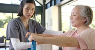 Asyalı kıdemli bir kadına direniş bandında yardım etmek, evde bakım sağlayan genç bir fizyoterapist. Fizik tedavi, üst düzey bakım, ev sağlığı, egzersiz, rehabilitasyon, sağlık hizmetleri