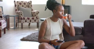 Afro-Amerikalı genç bir kadın oturmuş, çay yudumluyor, akıllı telefon tutuyor, evde. Saçları örgülü, beyaz atlet giyiyor ve açık renk hırka giyiyor, değişmemiş, ağır çekimde.