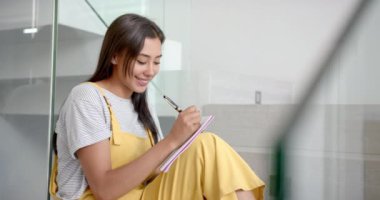 Gülümseyen kadın evinde oturmuş not defterine yazıyor, gününün tadını çıkarıyor, kopyalama alanı. Mutluluk, üretkenlik, ev ofisi, günlük, yaşam tarzı, yaratıcılık