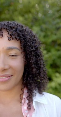 Dikey video: Kıvırcık saçlı gülümseyen kadın bahçede eğleniyor. Mutluluk, yaşam tarzı, doğa, rahatlama, yeşillik, rahatlık.