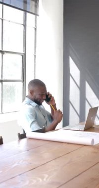 Dikey video: Akıllı telefondan konuşmak, adam dizüstü bilgisayar kullanıyor ve ofiste kalem tutuyor. İş, çoklu görev, teknoloji, iş, iletişim, meslek