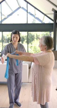 Dikey video: Fizyoterapist yaşlı kadına evde direnç bandı egzersizlerinde yardımcı oluyor. Fizyoterapi, rehabilitasyon, sağlık, fonksiyonel eğitim, fitness, terapi