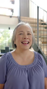 Dikey video: Mor bluzlu gülümseyen yaşlı kadın evdeki merdivenlerin önünde duruyor. Yaşlı, bağımsız, mutluluk, yaşam tarzı, günlük, kapalı