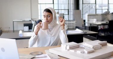 Kahve içen, tesettürlü kadın ofiste mimari model üzerinde çalışıyor. tasarım, çalışma alanı, konsantrasyon, yaratıcılık, kafein, üretkenlik
