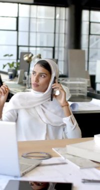 Dikey video: Akıllı telefondan konuşmak, tesettürlü kadın modern ofiste masa başında çalışmak. İş, işyeri, profesyonel, iletişim, teknoloji, üretkenlik
