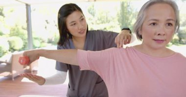 Kol egzersizi yapan yaşlı Asyalı bir kadına yardım etmek, evde dambıl kullanan bir fizyoterapist. Rehabilitasyon, fizyoterapi, sağlık hizmetleri, yaşlılar, fizik tedavi, dayanıklılık eğitimi