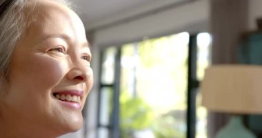 Gülümseyen kıdemli Asyalı kadın pencereden dışarı bakıyor, evde huzurlu bir anın tadını çıkarıyor. Mutluluk, memnuniyet, rahatlık, emeklilik, yaşam tarzı, kapalı alan
