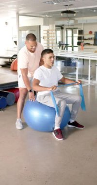 Dikey video: Direnç bandı egzersizlerinde yardımcı olmak, terapistin istikrar topunda adama yardım etmesi. Fitness, rehabilitasyon, fizyoterapi, fizik tedavi, egzersiz, eğitim, yavaş çekim