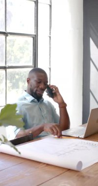 Dikey video: Telefonda konuşmak, adam ofiste dizüstü bilgisayarla mimari planları gözden geçiriyor. Mimarlık, iş, meslek, teknoloji, iletişim, çalışma alanı