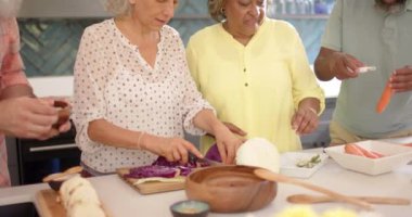 Bir grup kıdemli arkadaş mutfakta birlikte yemek hazırlıyor. Hepsinin gri saçları var ve günlük kıyafetler giyiyorlar, değiştirilmemiş, ağır çekimde.
