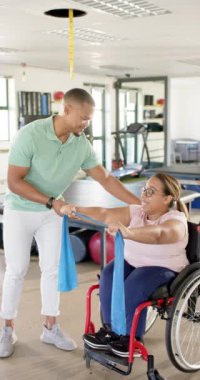 Dikey video: Tekerlekli sandalyedeki kadına direnç bandı egzersizlerinde yardımcı olan fizyoterapist. Fizyoterapi, rehabilitasyon, sağlık, sağlık, yardım, fitness, yavaş çekim