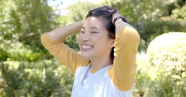 Sarı tişörtlü gülümseyen Asyalı kadın dışarıda eğleniyor, elleri saçında, şık giyiniyor. Mutluluk, doğa, yaşam tarzı, moda, gündelik, neşe