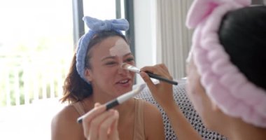 İki melez genç bayan arkadaş, yüz maskeleri takıp eğlenirken gülümsüyor. Geniş pencereli parlak oturma odası ve arka planda rahat mobilyalar, sıcak atmosfer yaratıyor, değişmemiş.