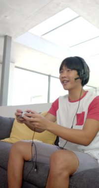 Dikey video oyunu oynamak, kulaklık takan Asyalı genç çocuk koltukta oturuyor. Oyun, eğlence, teknoloji, yaşam tarzı, eğlence, gençlik, yavaş çekim