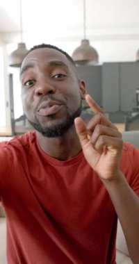 Dikey video: Kırmızı gömlekli Afro-Amerikalı adam evde otururken gülümsüyor ve el kol hareketi yapıyor. Mutluluk, kapalı alan, günlük, yaşam tarzı, neşeli, jest, yavaş çekim