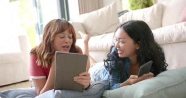 İki melez genç kız evde, bir tablet ve bir akıllı telefon kullanarak rahatlıyorlar. Büyük pencereli, değişmemiş, ağır çekimde, parlak, rahat oturma odasında, minderlerin üzerinde uzanarak.