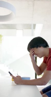 Dikey video: Asyalı genç çocuk kahvaltı yapıyor ve evde akıllı telefon kullanıyor, şaşırmış görünüyor. Dijital, sabah rutini, teknoloji, gündelik, yaşam tarzı, kapalı alan, sosyal medya, yavaş çekim