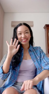 Dikey video: Ekose gömlek giyen gülümseyen Asyalı kadın, içeride oturuyor, kameraya bakıyor. Portre, gündelik, güven, yaşam tarzı, moda, yavaş çekim