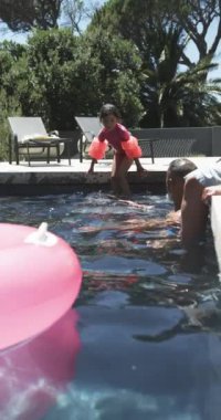 Dikey video: Havuz kenarında oturan şamandıralı çocuk, yüzme etkinliğine yardım eden büyükanne ve büyükbaba. aile, yaz, tatil, su, güvenlik, eğlence