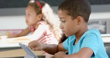 Çift ırklı erkek ve kız okuldaki tabletlerine odaklandılar. Bir masada otururlar, eğitim faaliyetleri veya oyunlarla meşguldürler, yavaş çekimde..
