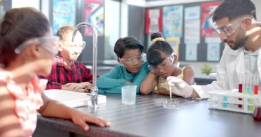 Çeşitli bir grup genç öğrenci bilim deneyi yapıyor. Siyah saçlı ve gözlüklü bir Asyalı öğretmen onlara önderlik ediyor. Hepsi koruyucu gözlük takıyor.