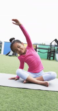 Dikey video: Okul bahçesinde, melez bir kız yoga yapıyor. Saçlarını bağlamış, pembe bir bluz ve açık mavi taytlar giyiyor, duruşuna odaklanıyor..