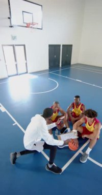 Dikey video: Basketbol koçu sahada oturan erkek oyuncularla strateji tartışıyor. Koçluk, takım çalışması, spor, antrenman, toplanma, atletizm