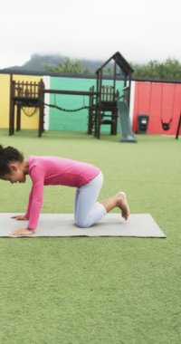 Dikey video: Okul parkında, genç bir kız minder üzerinde yoga yapıyor. Asyalı, siyah saçlı, pembe bluzlu ve beyaz taytlı.
