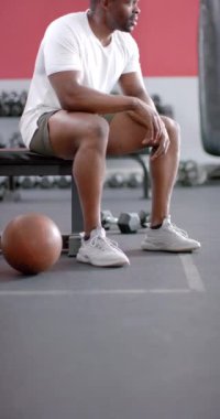 Dikey video: yedek kulübesinde dinlenen, güçlü ve formda bir adam spor salonunda terleyerek yakınlarda basketbol oynuyor. Spor, spor, egzersiz, antrenman, spor, ağır çekim
