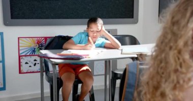 Çift ırklı çocuk elinde kalemle ve masada kitaplarla sınıfta yazmaya odaklanmış. Güneş ışığı odayı dolduruyor, renkli duvar sanatı ve eğitim ortamını vurguluyor..