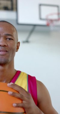 Dikey video: Basketbol topu tutmak, spor formalı gülümseyen adam kapalı sahada duruyor. Sporcu, kendine güven, aktif, rekabet, hobi, coşku
