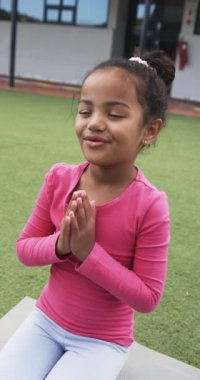 Dikey video: Okul ortamında, melez genç kız bacak bacak üstüne atmış oturuyor. Gözleri kapalı, elleri bir arada ve huzur içinde meditasyon yapıyor gibi görünüyor.