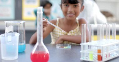 Okulun kimya laboratuvarında, melez bir öğrenci gülümsüyor. Arkasında, melez bir öğretmen posterlerin ve camların arasında, değiştirilmemiş, ağır çekimde denetliyor..