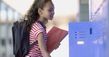 Kıvırcık saçlı melez kız okul dolaplarının yanında duruyor. Çizgili bir gömlek ve sırt çantası giyiyor, tipik bir öğrenci gününü temsil ediyor, ağır çekimde..