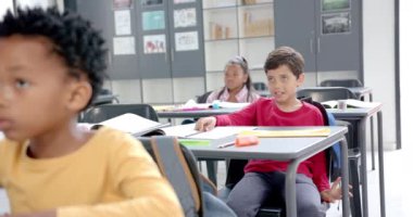 Okulda, çocuklar sınıflarında renkli malzemelerle masalarında oturuyorlardı. Bir çocuk arkadaşıyla konuşuyor. Etrafı tablolar ve matematik işaretleriyle çevrili. Değiştirilmemiş, ağır çekim..