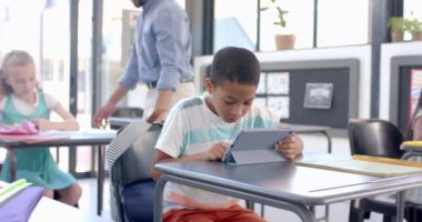 Çift ırklı çocuk okuldaki bir tablete odaklanmış. Bir öğretmen parlak, modern bir sınıf ortamında öğrencilere yardımcı olur, yavaş çekimde..