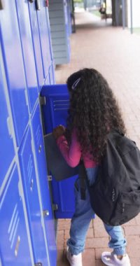 Dikey video: Okul koridorunda, dolabında melez bir kız var. Kıvırcık siyah saçları var, günlük kıyafetler giyiyor ve elinde mavi bir dosya var..