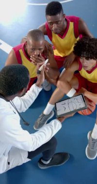 Dikey video: Basketbol koçu sahadaki erkek oyunculara stratejiyi açıklamak için tablet kullanıyor. Spor, takım çalışması, koçluk, eğitim, teknoloji, tartışma