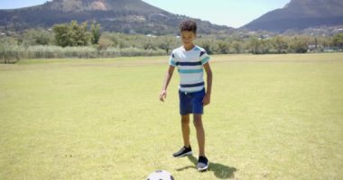 Kıvırcık saçlı melez çocuk okuldaki güneşli bir parkta ayağının dibinde futbol topuyla gülümsüyor. Çizgili bir tişört ve şort giyiyor, gençlik enerjisi yayıyor ve sporu seviyor..